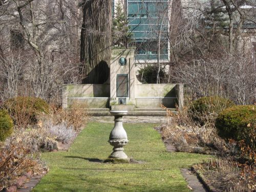 A serene garden scene featuring a stone fountain, manicured hedges, and a backdrop of trees and a modern building.