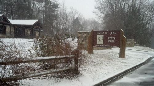 Sign for Caesars Head State Park in a snowy landscape, with a rustic building and trees in the background.