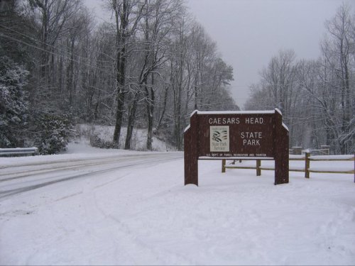 Snow-covered entrance sign for Caesars Head State Park, surrounded by bare trees and a snowy road.