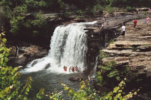 A waterfall cascades into a pool, with people swimming and relaxing on the rocks nearby, surrounded by lush greenery.