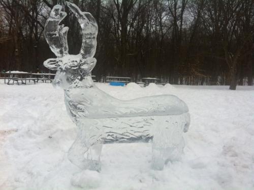 A large ice sculpture of a deer stands in a snowy landscape, surrounded by trees and picnic tables.