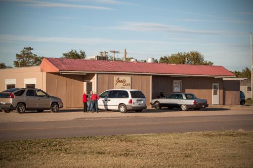 A small café with a red roof, parked cars, and people chatting outside on a sunny day.