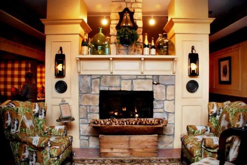 Cozy living room with a stone fireplace, two patterned chairs, and decorative bottles on the mantel. Warm and inviting atmosphere.