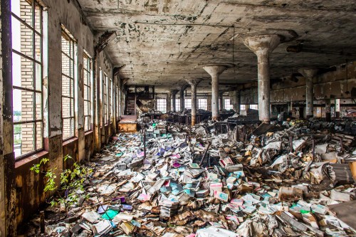 An abandoned, dilapidated room filled with scattered books and debris, with large windows letting in natural light.
