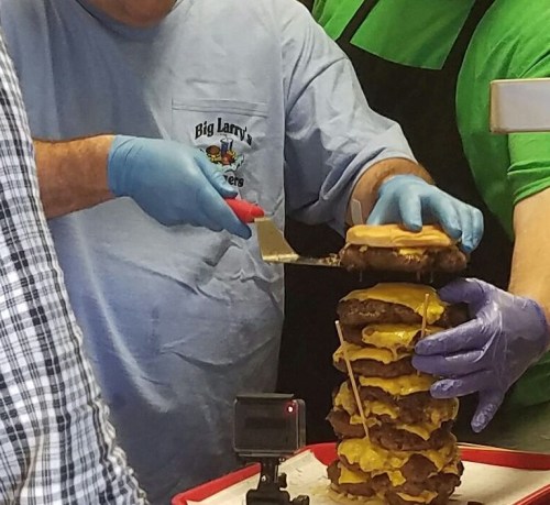 A person in a blue shirt and gloves is stacking a towering burger with multiple patties and cheese slices.