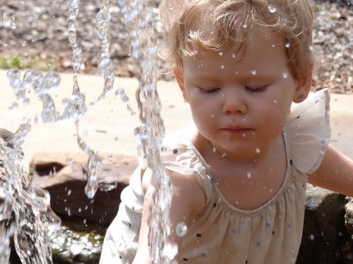 A young child with curly hair plays near a water fountain, eyes closed, surrounded by sparkling droplets.