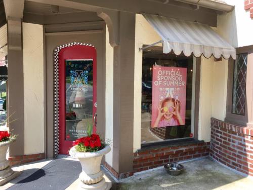Exterior of a storefront with a red door, awning, and a sign reading "Official Sponsor of Summer." Flower pots in front.