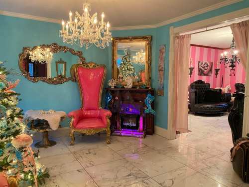 A vibrant room with a pink throne chair, chandeliers, and a decorated Christmas tree, featuring colorful walls and elegant decor.