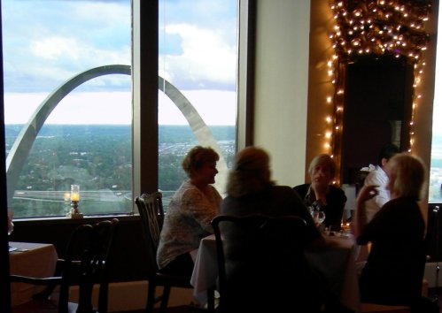 A group of women dining in a restaurant with a view of a large arch structure outside the window.
