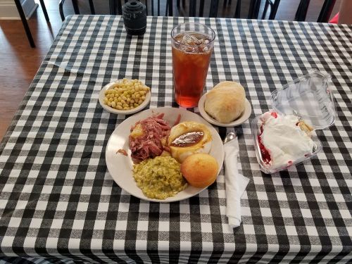 A plate of pulled pork, green beans, cornbread, and a roll, with a side of corn and a dessert, on a checkered tablecloth.