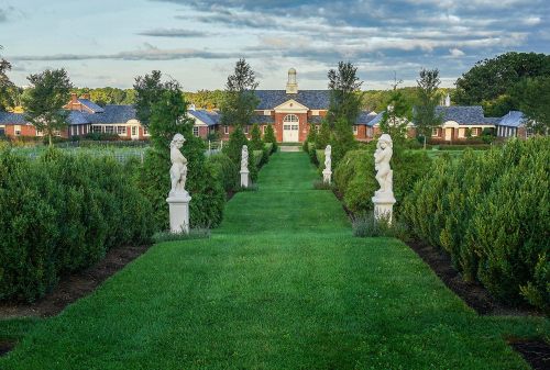 A manicured garden path lined with statues leads to a brick building under a cloudy sky.