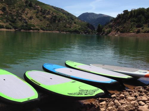 Colorful paddleboards lined up on a rocky shore by a calm lake, surrounded by green hills and blue skies.