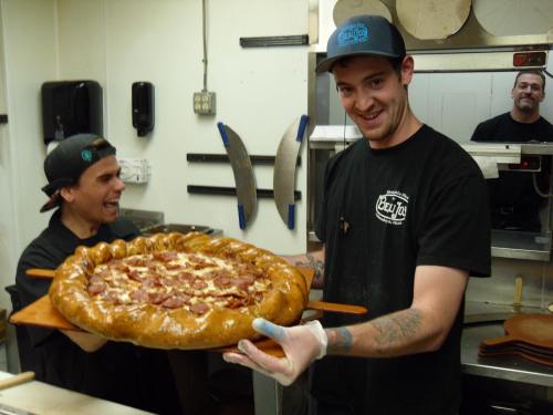 Two chefs in a kitchen proudly hold a large, uniquely shaped pizza with pepperoni, smiling at the camera.