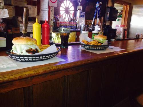Two baskets of burgers with lettuce and fries on a wooden bar, alongside condiments and a glass of soda.