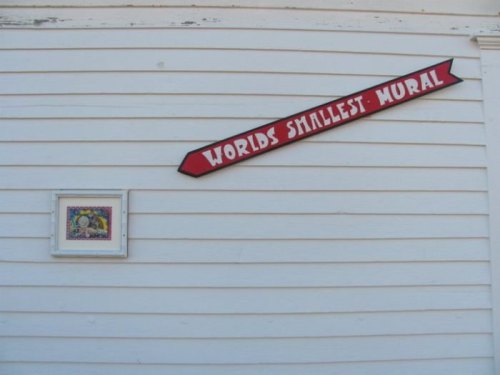 Sign reading "World's Smallest Mural" next to a small framed mural on a white wall.