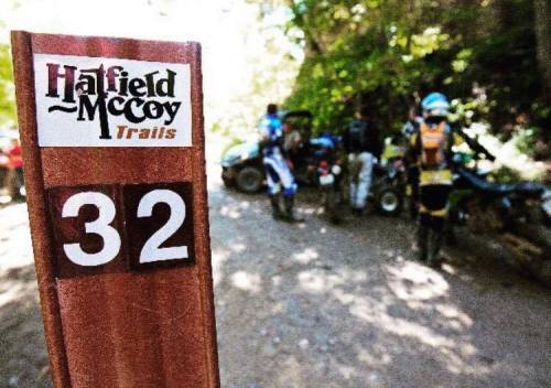 Signpost for Hatfield-McCoy Trails, marked 32, with riders and vehicles in the background on a wooded trail.