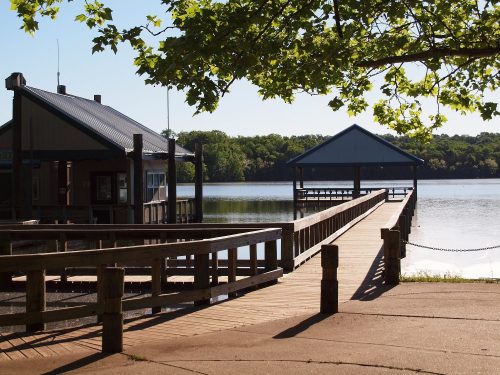 A wooden dock extends over calm water, with a small building and gazebo nearby, surrounded by trees.