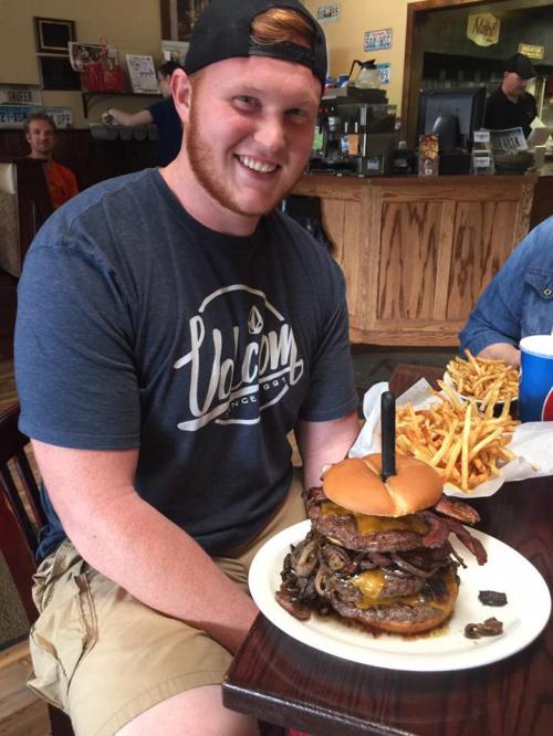 A smiling man sits at a table with a massive burger topped with bacon and cheese, surrounded by fries.