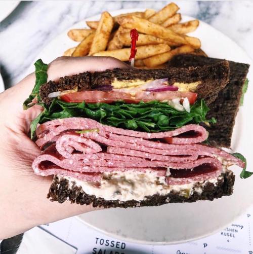 A hand holding a large sandwich with layers of meat, lettuce, tomato, and mustard, alongside a plate of fries.