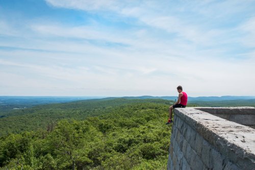 A person sits on a stone ledge overlooking a lush green landscape and distant mountains under a blue sky.