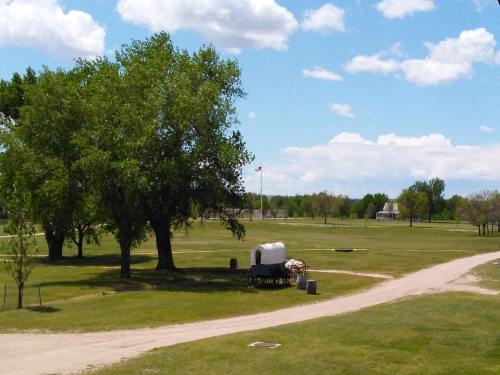 A grassy landscape with a covered wagon, trees, and a distant flag under a blue sky with fluffy clouds.