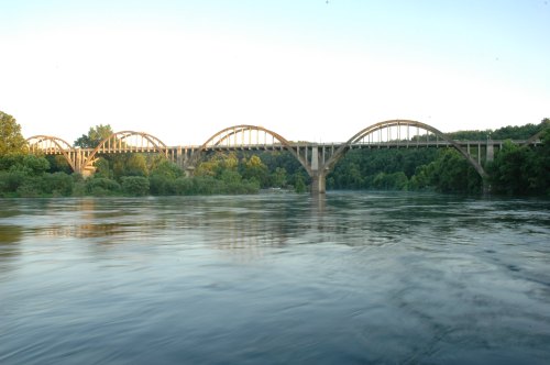 A large arched bridge spans a calm river, surrounded by lush green trees under a clear sky.