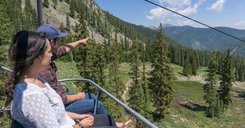 A man points out a view while riding a chairlift with a woman, surrounded by trees and mountains on a sunny day.