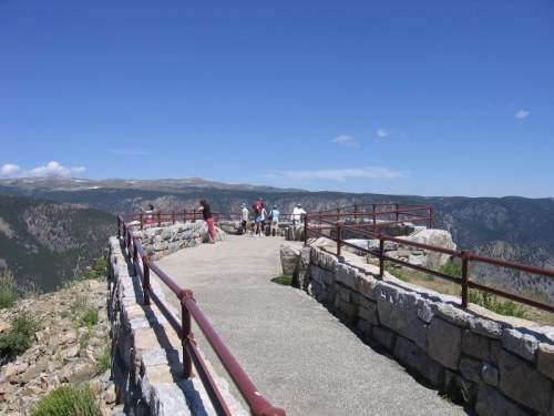 A scenic overlook with a stone pathway and railing, where several people enjoy the view of mountains and blue sky.