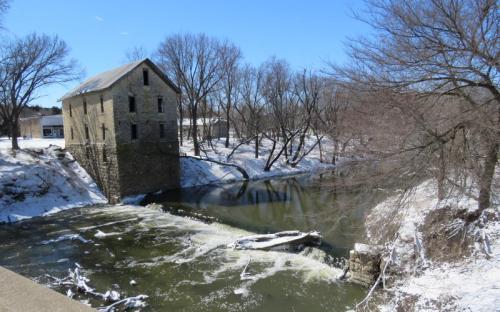 A stone building by a snowy river, with bare trees and a clear blue sky in the background.