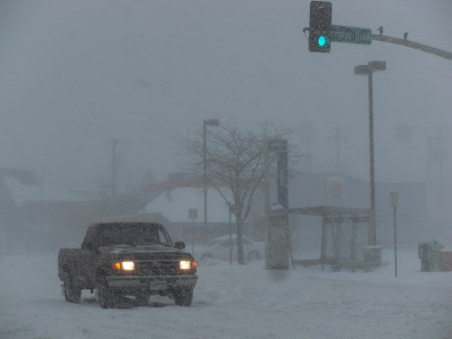 A truck navigates a snowy street with low visibility, near a traffic light and a bus stop in a winter storm.