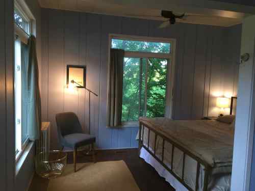 Cozy bedroom with a bed, chair, and large window overlooking greenery, featuring soft lighting and blue walls.