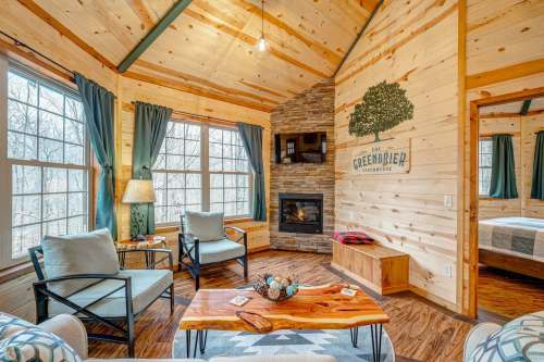 Cozy cabin interior with wooden walls, a stone fireplace, and large windows, featuring comfortable seating and decor.