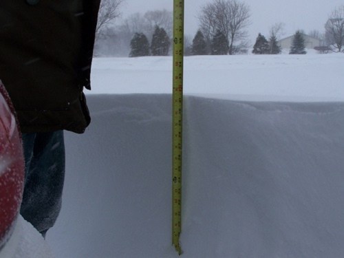 A measuring tape stands upright in deep snow, showing significant snowfall accumulation during a winter storm.