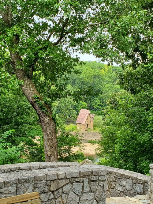 A rustic stone building with a red roof surrounded by lush greenery and trees, viewed from a stone wall.