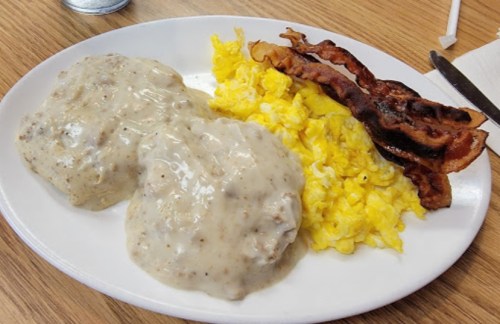 A plate of biscuits and gravy, scrambled eggs, and crispy bacon on a wooden table.