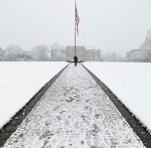 A person stands on a snowy path in front of a large building, with an American flag flying in the background.