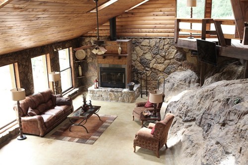 Cozy living room with stone walls, wooden beams, a fireplace, and comfortable seating. Natural light floods the space.