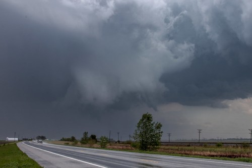 Dark storm clouds loom over a highway, with a hint of a funnel shape forming in the sky. Trees line the roadside.
