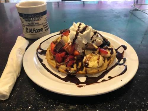 A plate of waffles topped with strawberries, bananas, whipped cream, chocolate drizzle, and a coffee cup in the background.