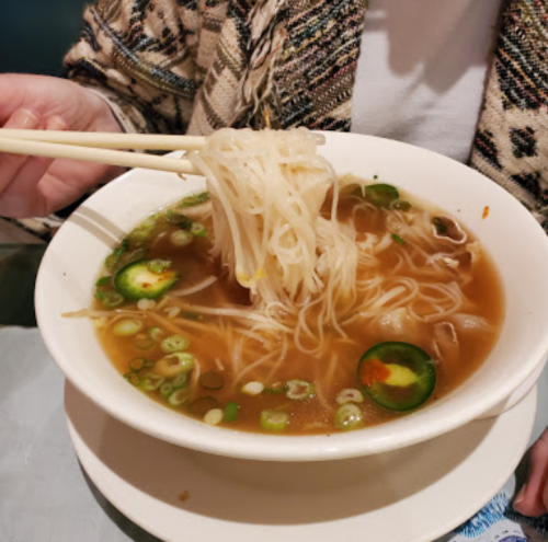 A bowl of noodle soup with green onions and jalapeños, held with chopsticks.