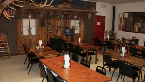 A rustic dining area with wooden tables, black chairs, and a wall decorated with antlers and a cabin-like mural.