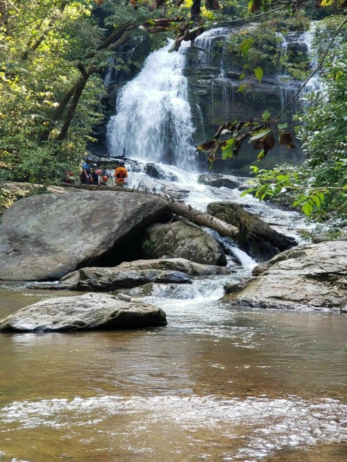 A scenic waterfall cascades down rocky cliffs, surrounded by lush greenery and a calm river with people in the background.