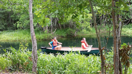 Two people in life jackets sit in a canoe on a calm, green river surrounded by lush vegetation.