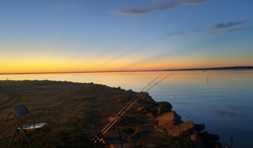 A serene lakeside view at sunset, with fishing rods set up and a chair nearby, reflecting calm waters and colorful skies.