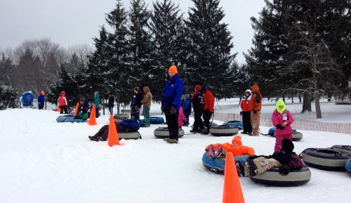 People in winter clothing enjoy snow tubing on a snowy hill, with orange cones marking the area and trees in the background.