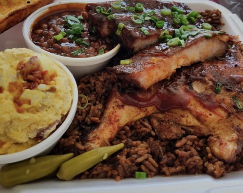 A plate of barbecue ribs, chicken, rice, beans, and mashed potatoes, garnished with green onions and pickles.