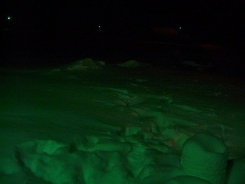 A snowy landscape illuminated by green light, with mounds of snow and faint distant lights visible in the background.