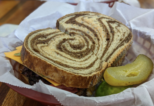 A sandwich on marbled rye bread with cheese and pickles, served in a basket lined with paper.