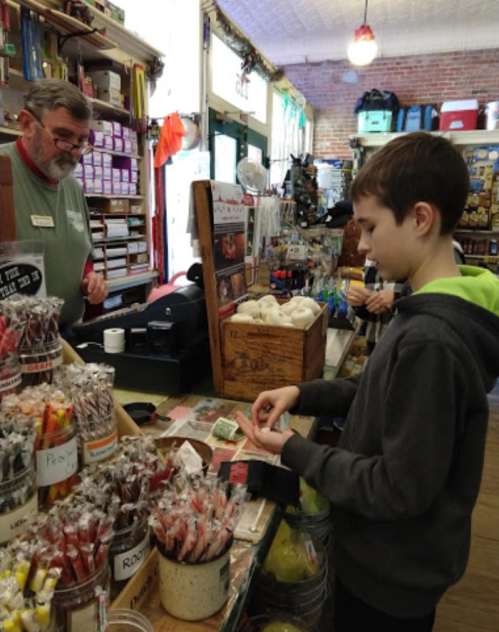A boy examines candy at a shop counter while an older man assists him. Colorful treats are displayed around them.