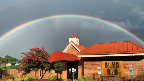 A vibrant rainbow arcs over a brick building with a red roof, surrounded by greenery and flowers.
